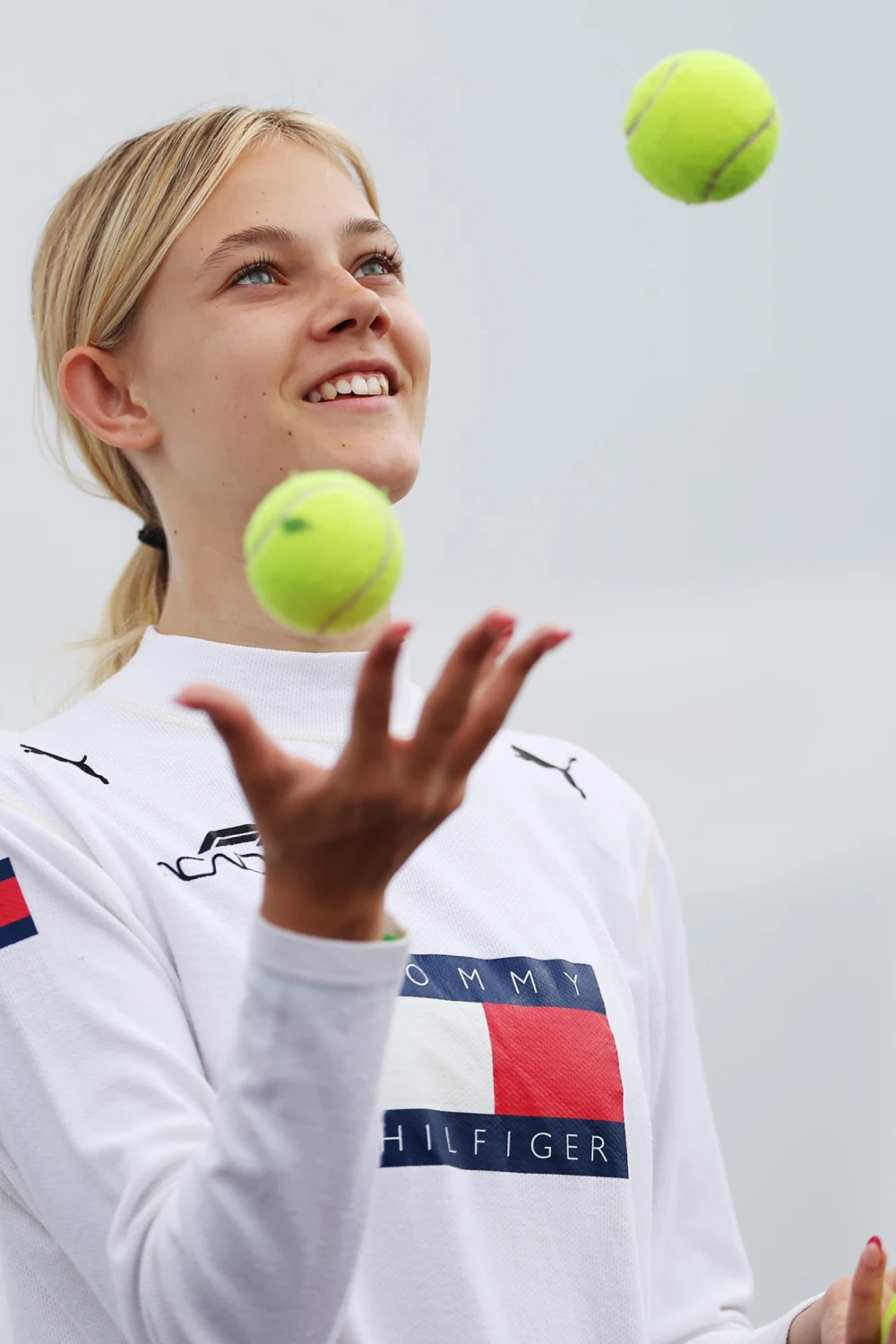 Young woman with blonde hair smiling and juggling two tennis balls against a gray sky.